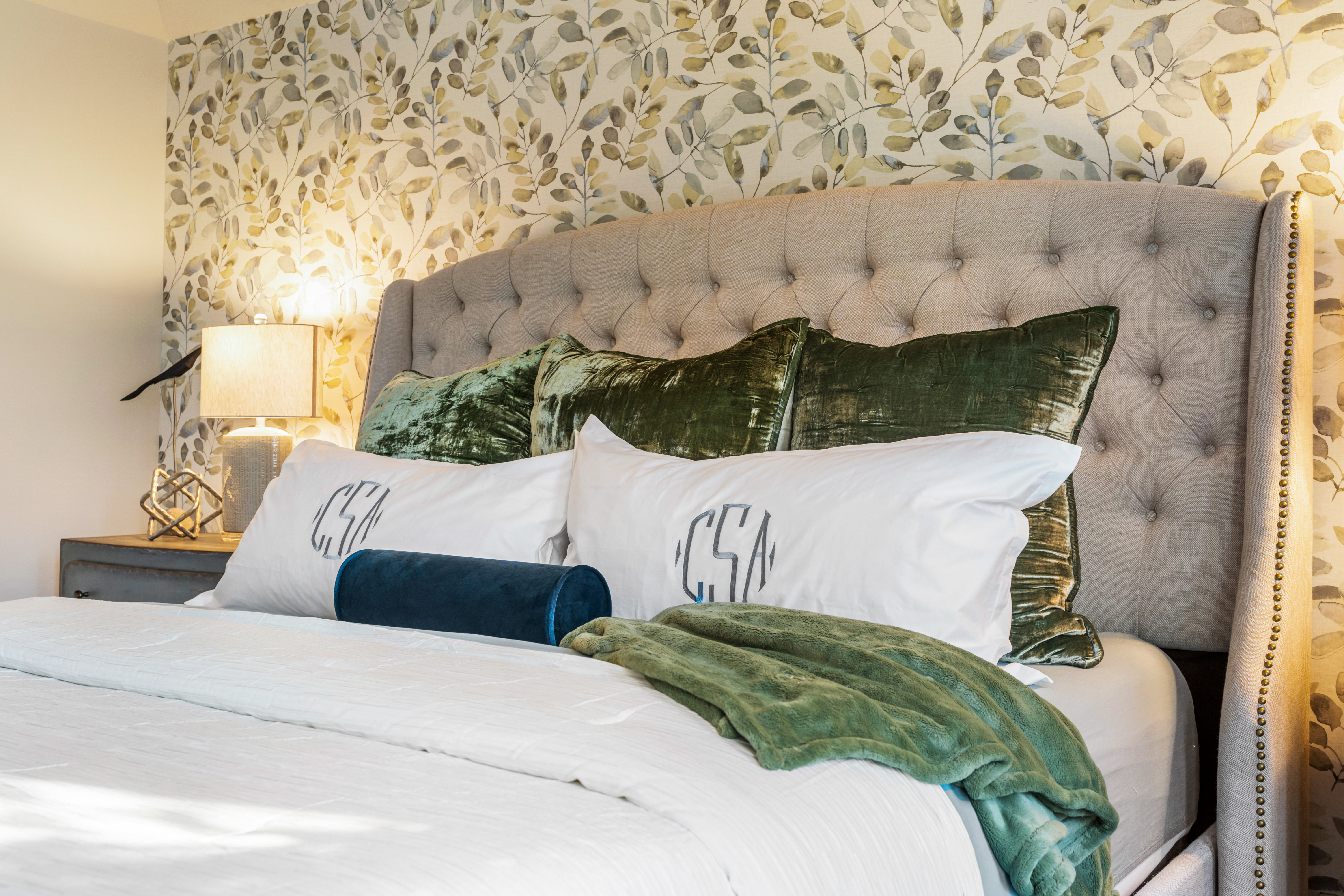 bed with green pillows and tan headboard against a green leaf wallpapered wall